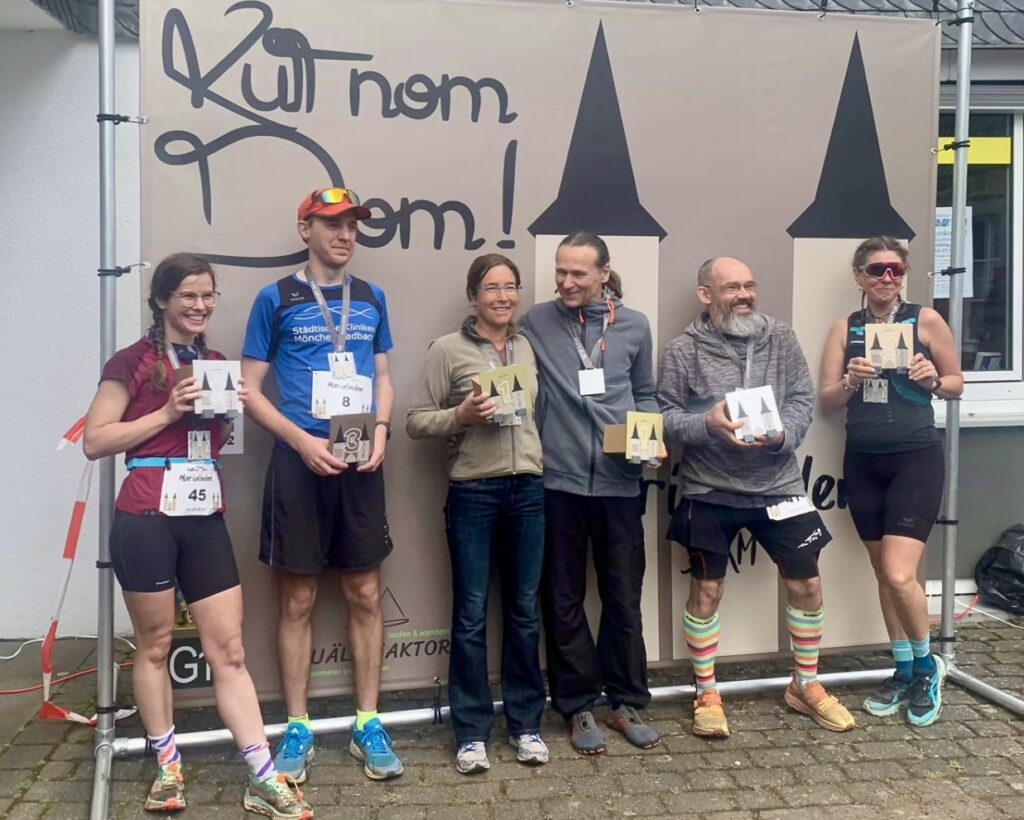 Personen vor einem Banner mit dem Titel "Kutt nom Dom" und einer Kirche auf dem Banner. Personen halten Pokale in der Hand.